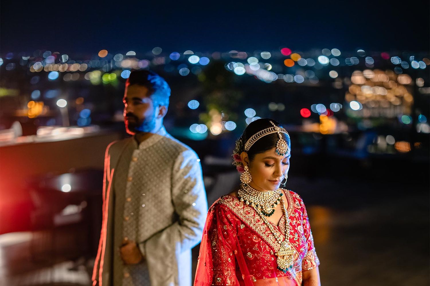 A couple in traditional Indian wedding attire stands against a nighttime city skyline with glowing bokeh lights.