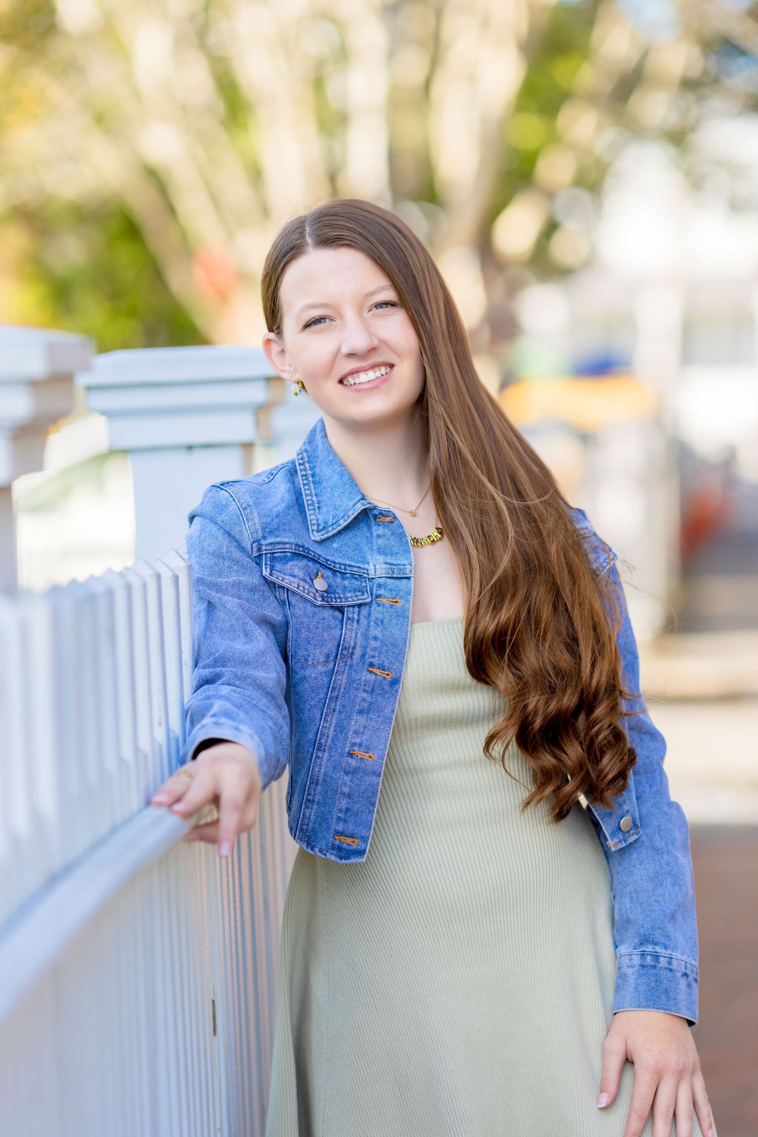 Young person wearing a denim jacket and green dress poses outdoors by a white railing on a sunny day.