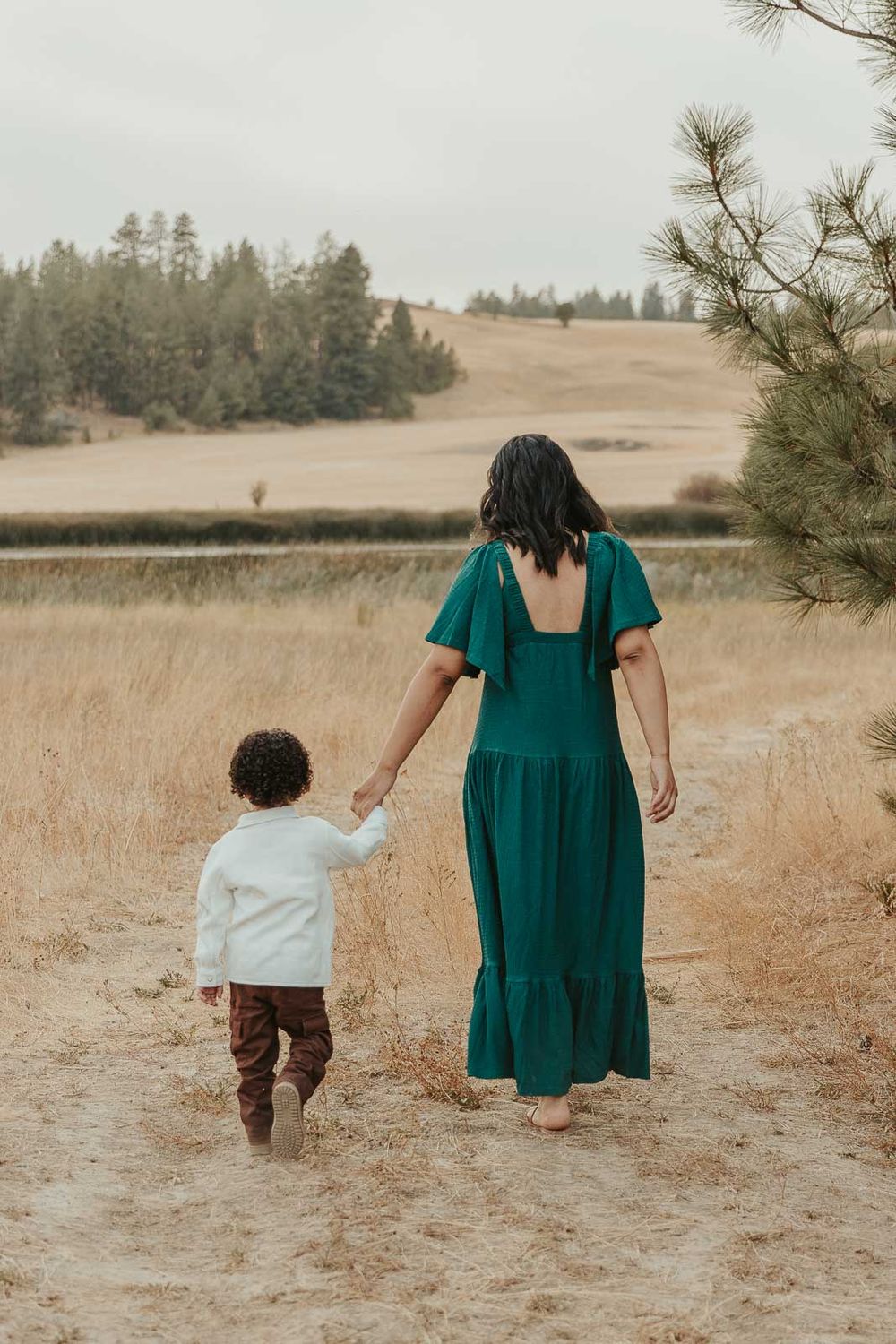 Expecting mother cradling her baby bump and walking with toddler son in a deep green maternity dress, surrounded by nature in Spokane