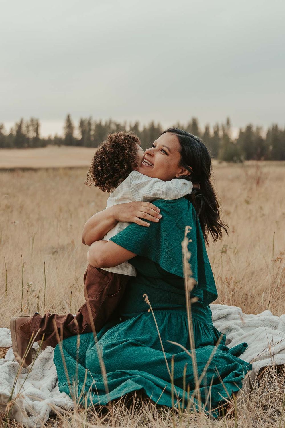 Expecting mother cradling her baby bump and hugging toddler son in a deep green maternity dress, surrounded by nature in Spokane