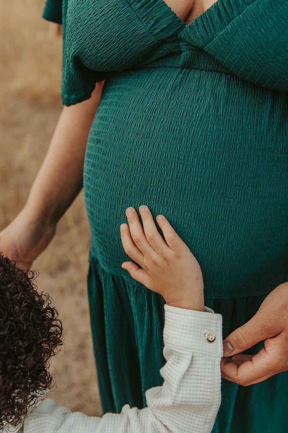 Expecting mother cradling and son's hand on baby bump in a deep green maternity dress, surrounded by nature in Spokane