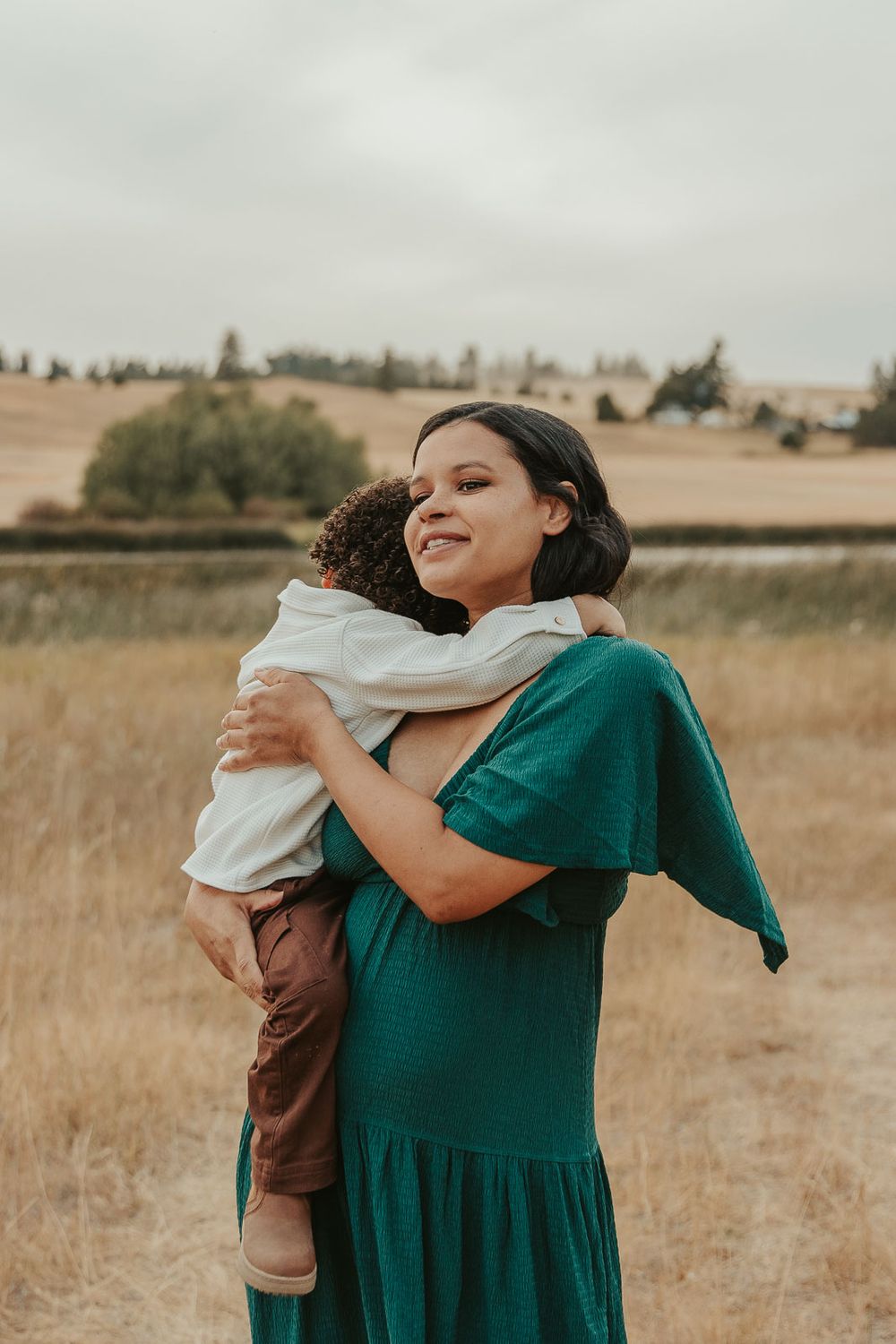 Expecting mother cradling her baby bump and snuggling toddler son in a deep green maternity dress in nature in Spokane, Spokane photographer