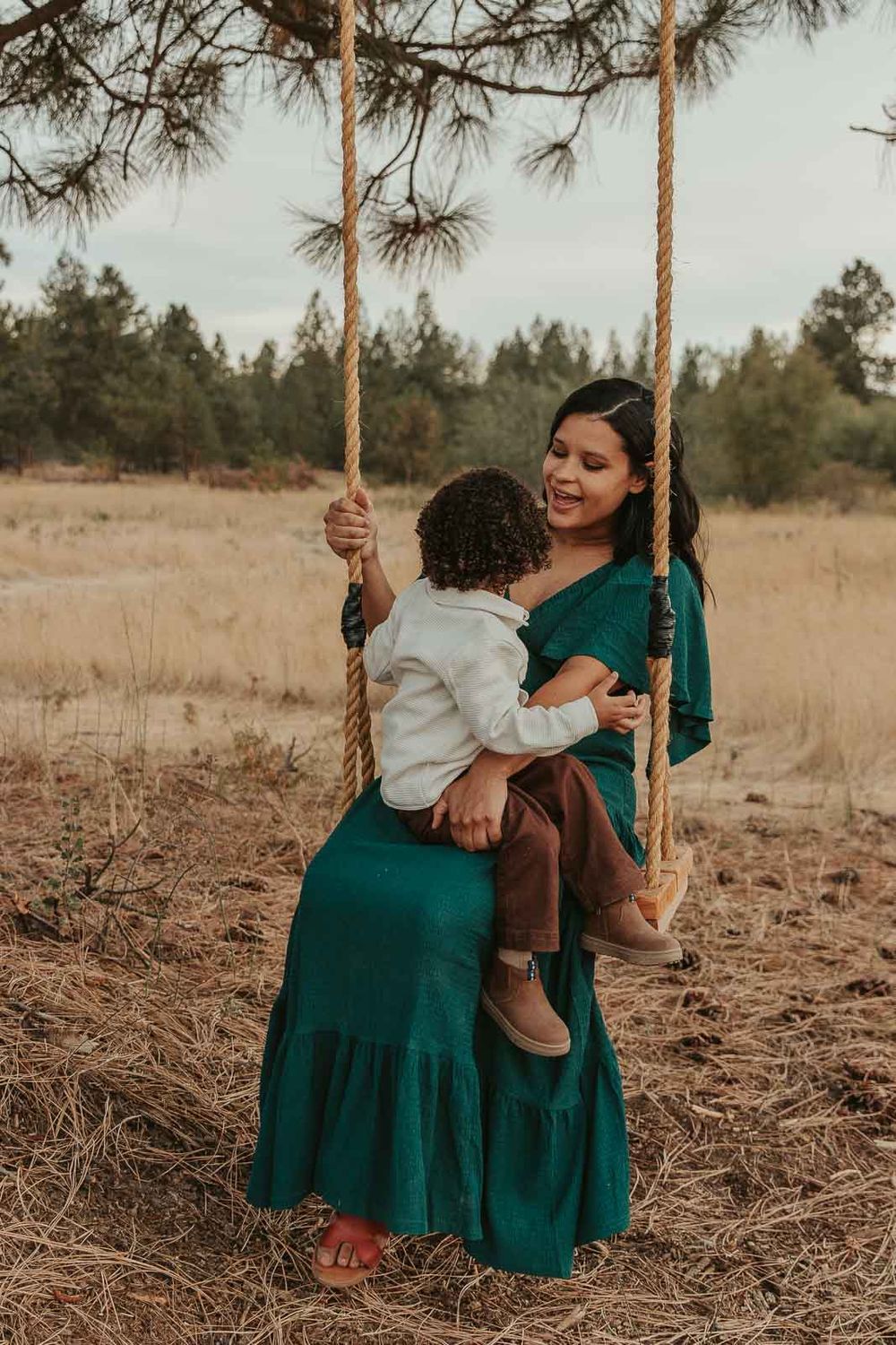 Expecting mother cradling her baby bump and pushing toddler son in tree swing in a deep green maternity dress, in nature in Spokane
