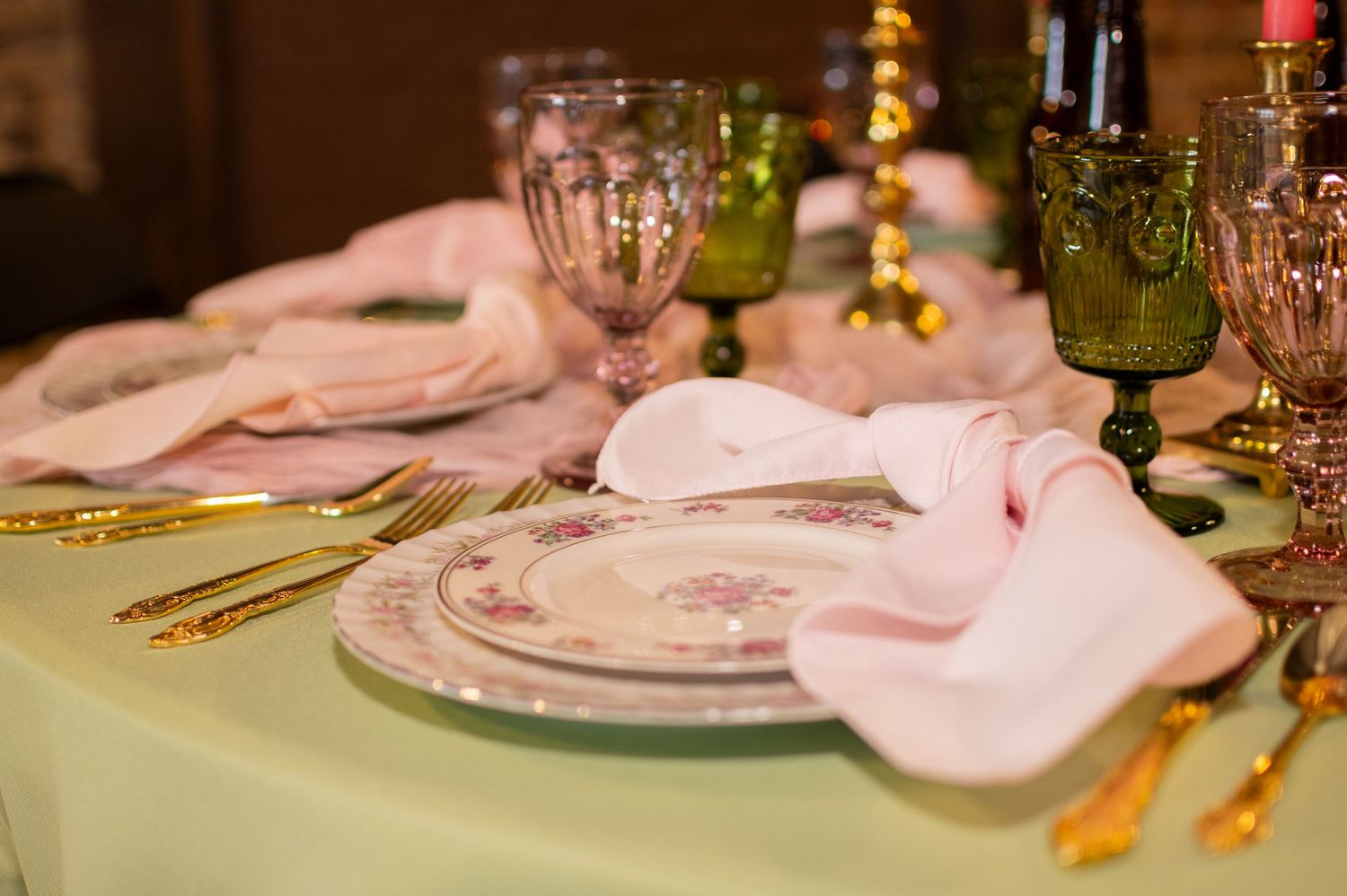 An elegant table setting with green glassware, pink napkins and gold cutlery on a light green tablecloth.