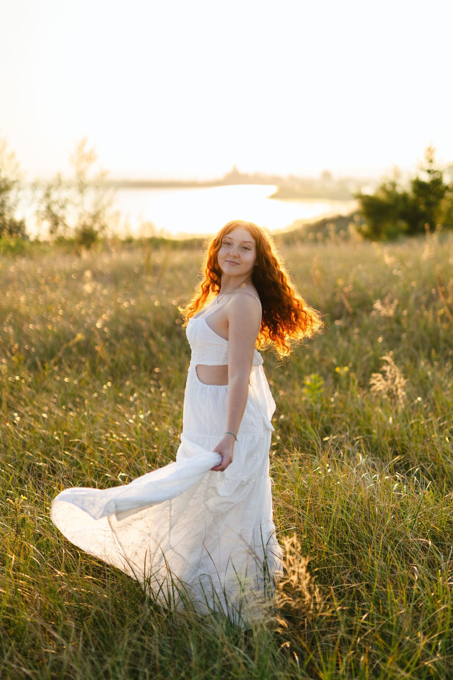 Woman in flowing white dress twirls in golden sunlit meadow at sunset by the water.