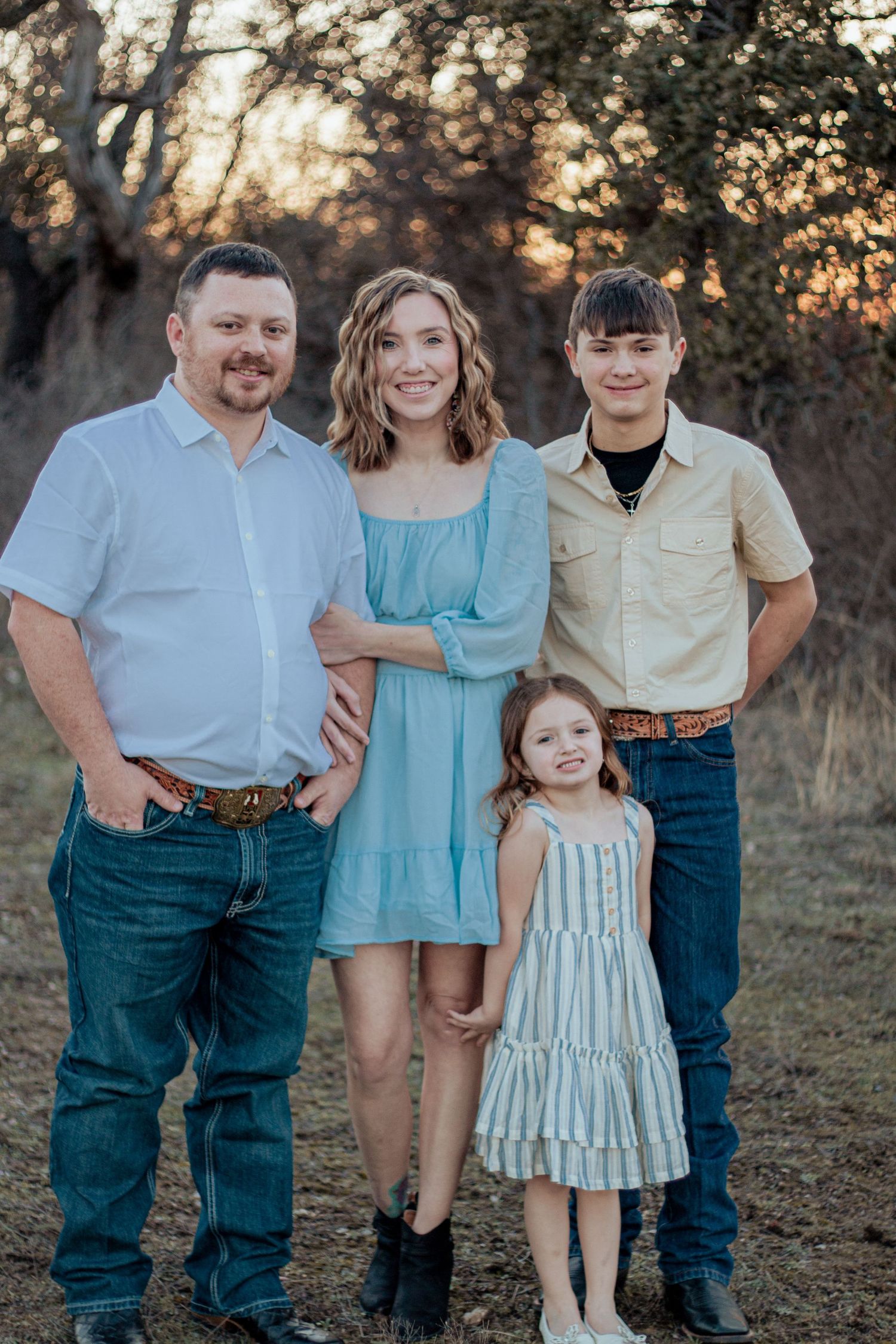 A family of four poses together outdoors at sunset wearing coordinating casual outfits in blue and neutral tones.
