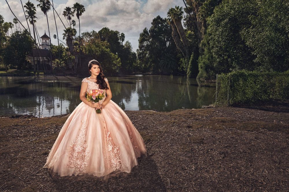 A stunning peach quinceañera ball gown with lace details photographed at sunset by a serene lake surrounded by palm trees.