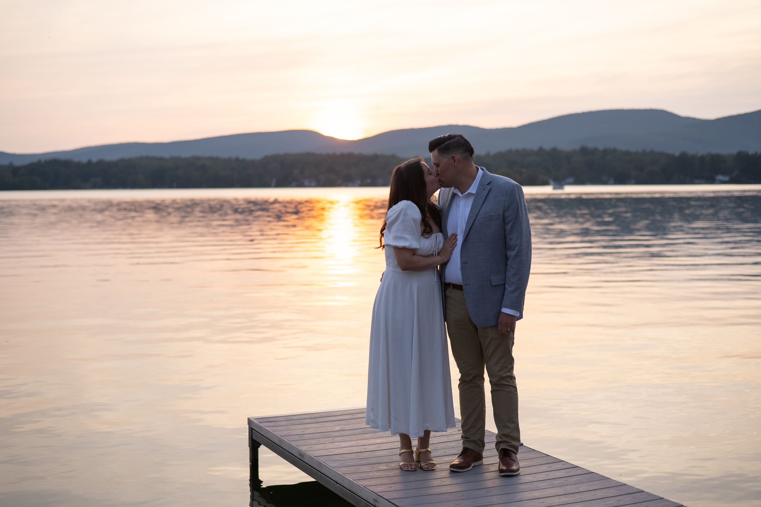 A couple stands embracing on a wooden dock at sunset with mountains and a tranquil lake in the background.