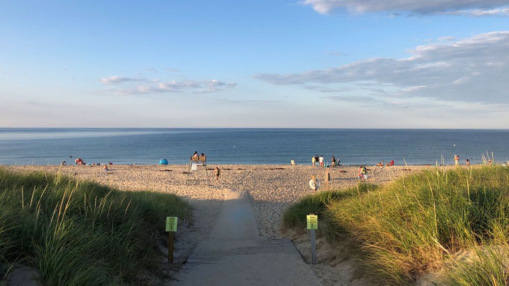 A sandy pathway leading through dunes to a beach with people visible in the distance.