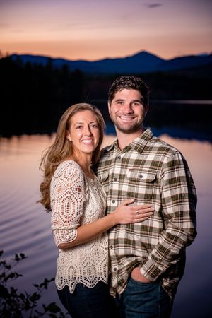 Couple poses by serene lake at sunset with mountains and forest in background.