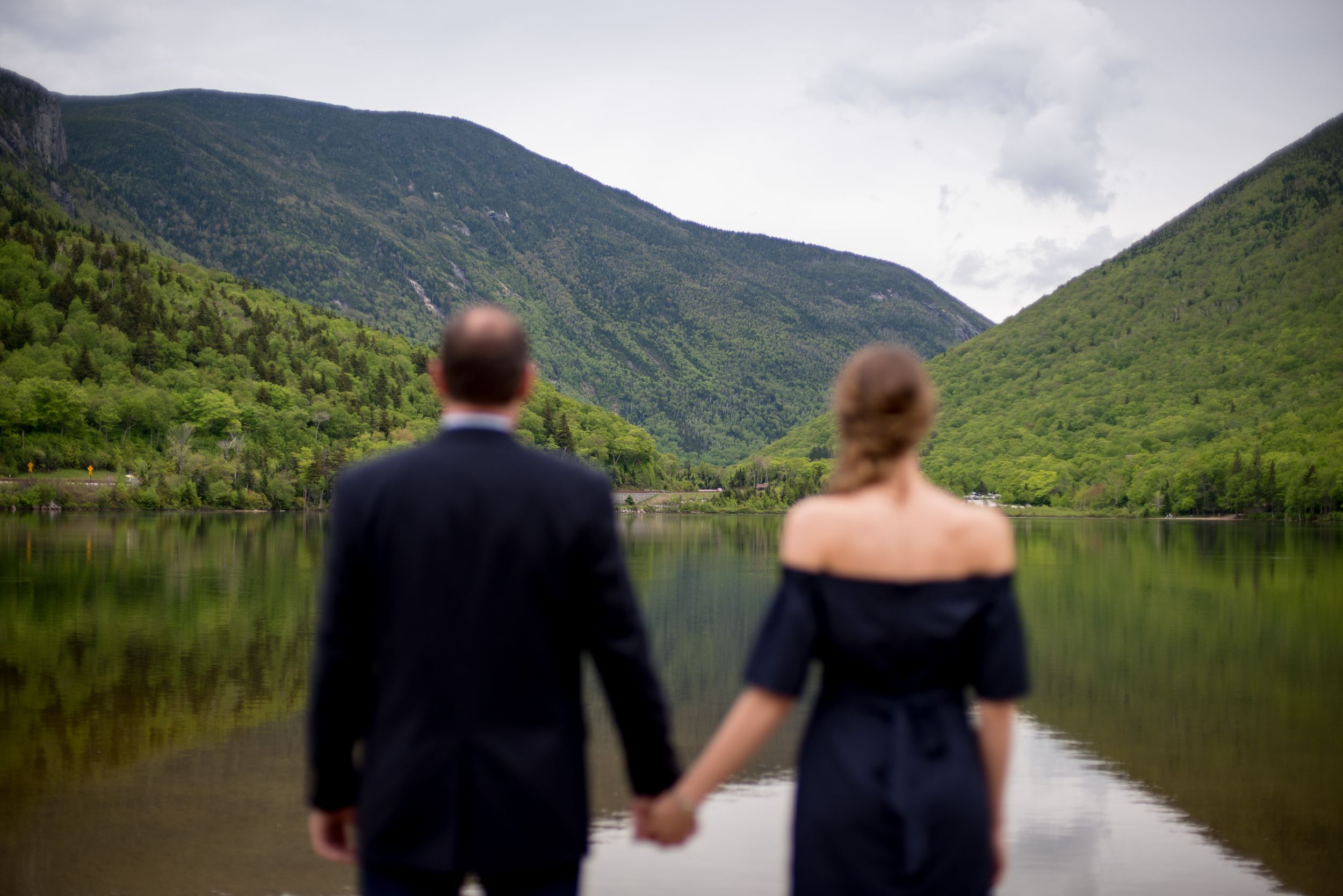 Two people stand together overlooking a peaceful mountain lake surrounded by green hills.