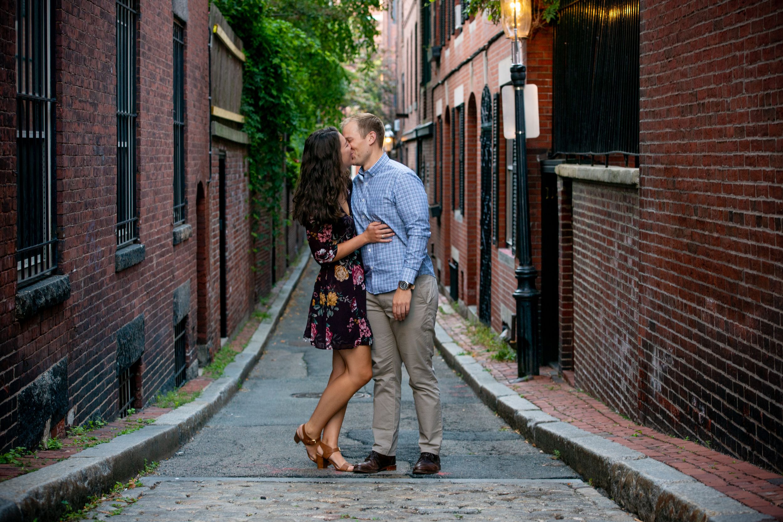 Two people walk along a historic cobblestone street lined with red brick buildings.