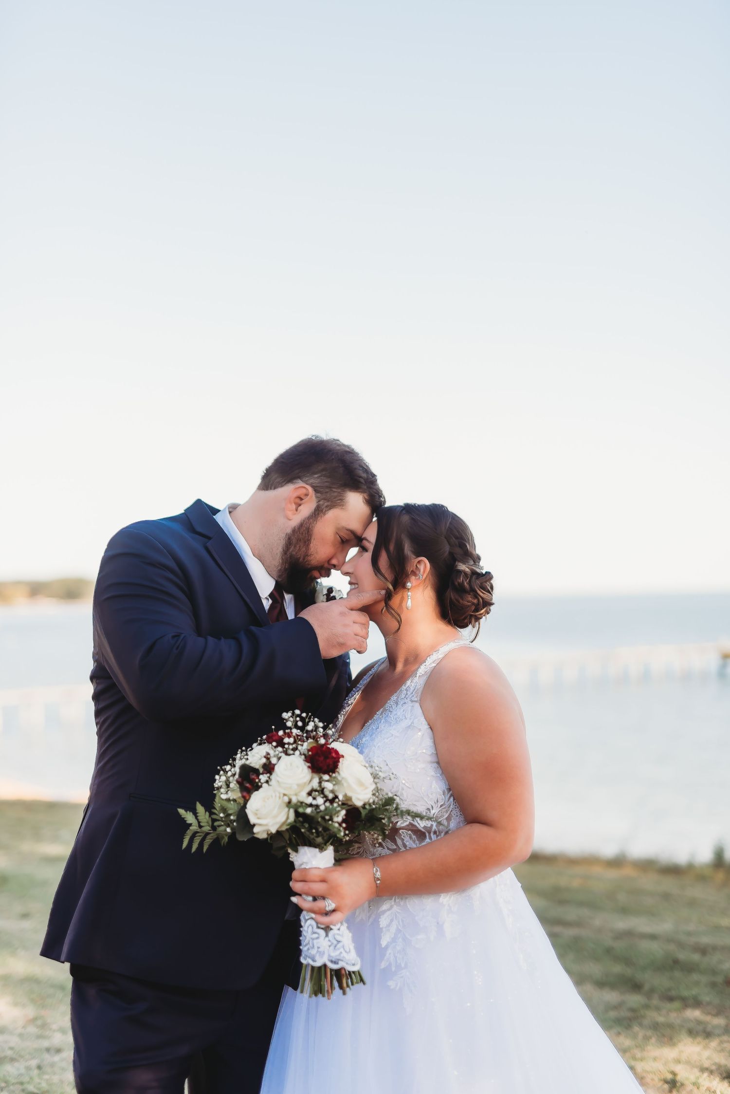 A romantic seaside wedding portrait with a couple sharing an intimate moment while holding a white and burgundy bouquet.