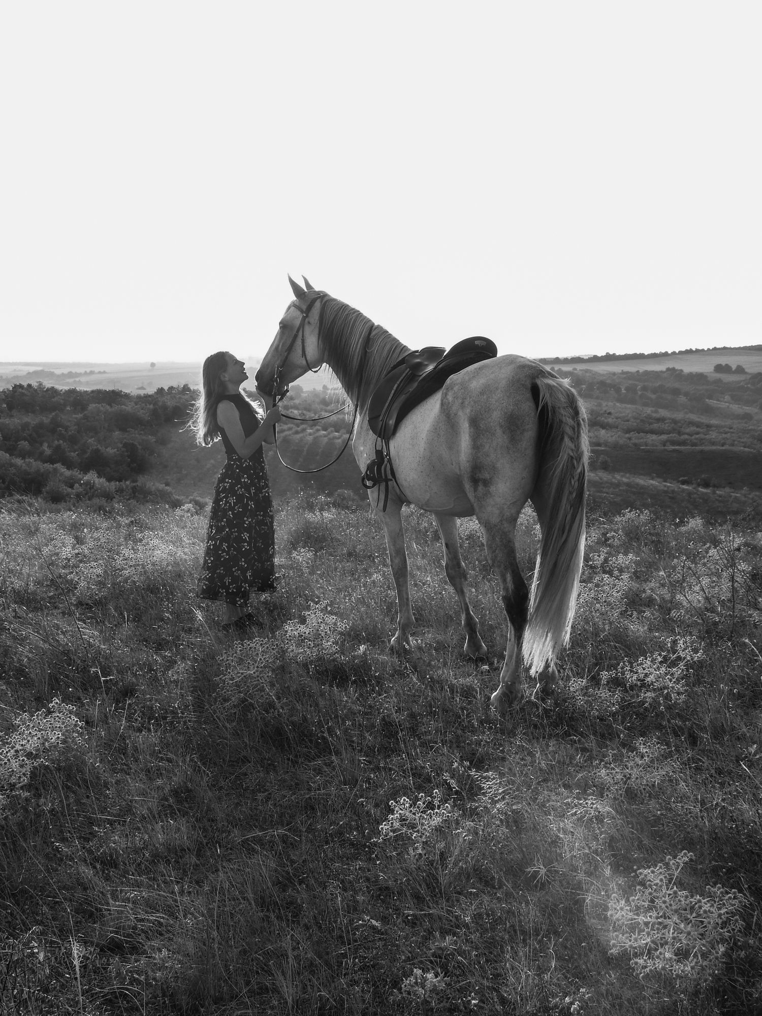 Black and white photo of person in dress standing beside horse in countryside.