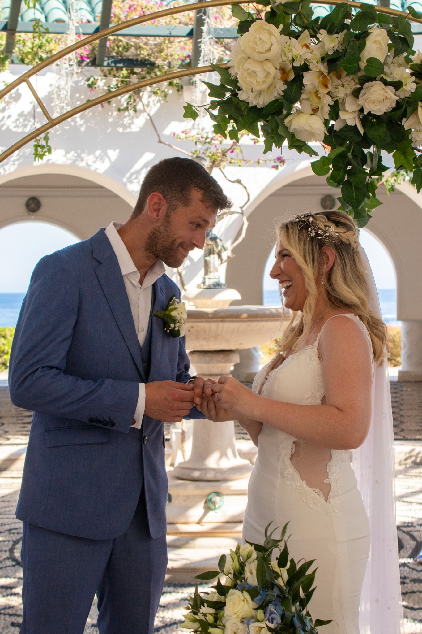 Wedding ceremony under floral arch with couple exchanging rings by the ocean.