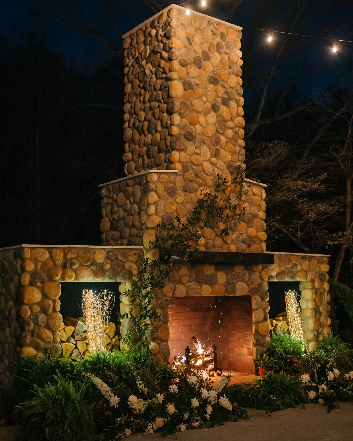 Beautiful stone outdoor fireplace illuminated at night with greenery and string lights.