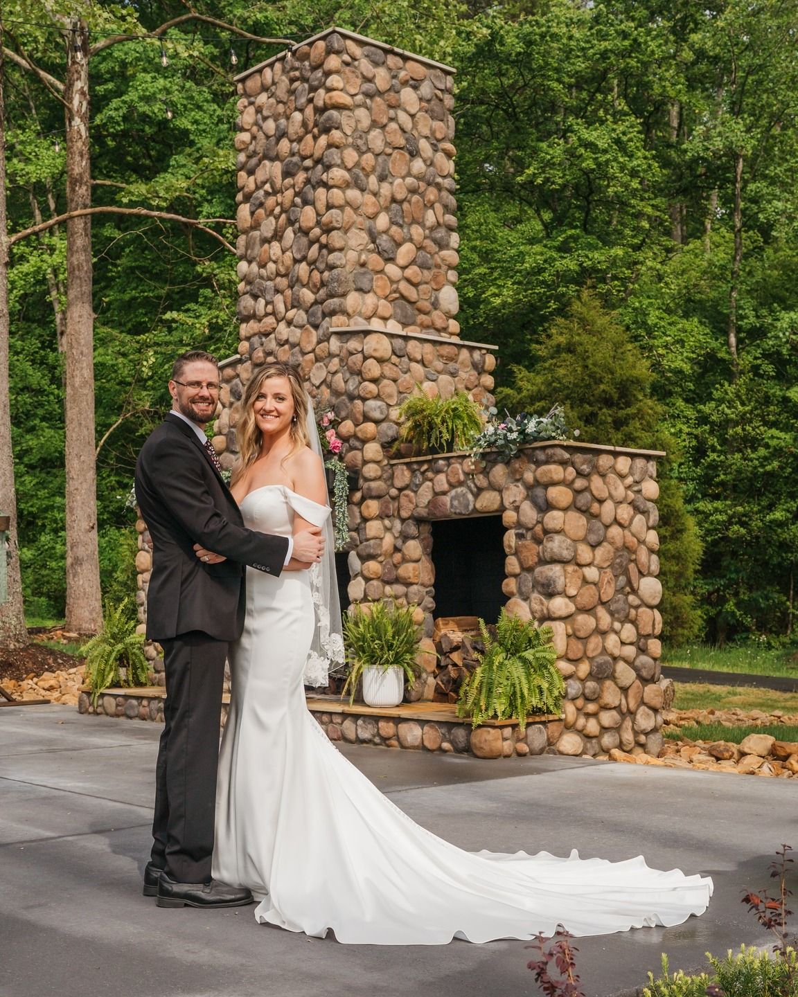 A couple poses by a rustic stone fireplace outdoors during a wedding celebration.