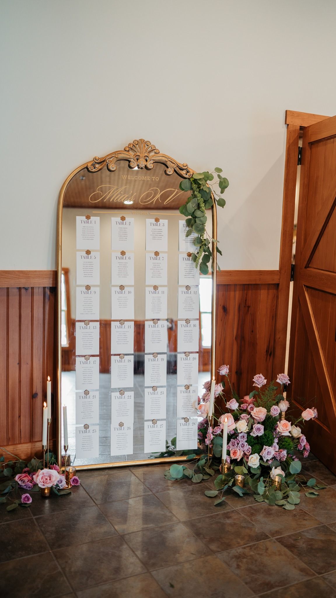 An elegant gold-framed mirror seating chart display adorned with pink roses and greenery.