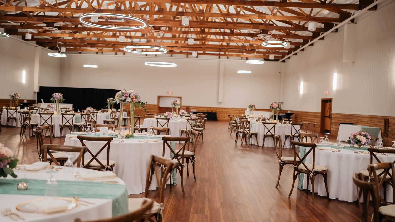 A spacious wedding reception venue with round tables, cross-back chairs, and rustic wooden beam ceiling.