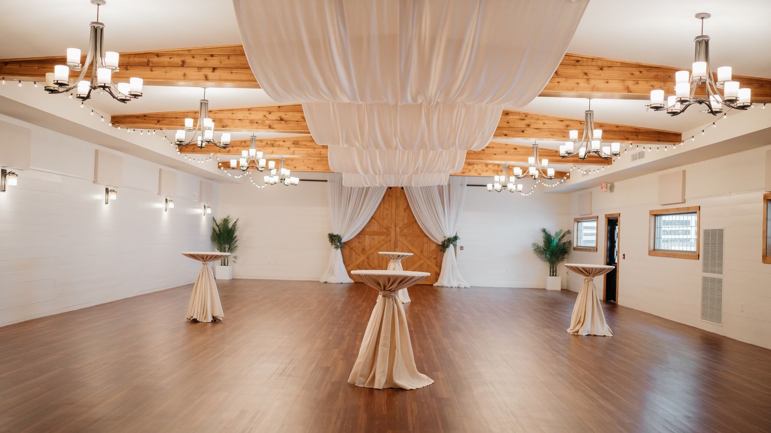 An elegant wedding reception space with draped fabric ceiling, chandeliers and wooden beams.