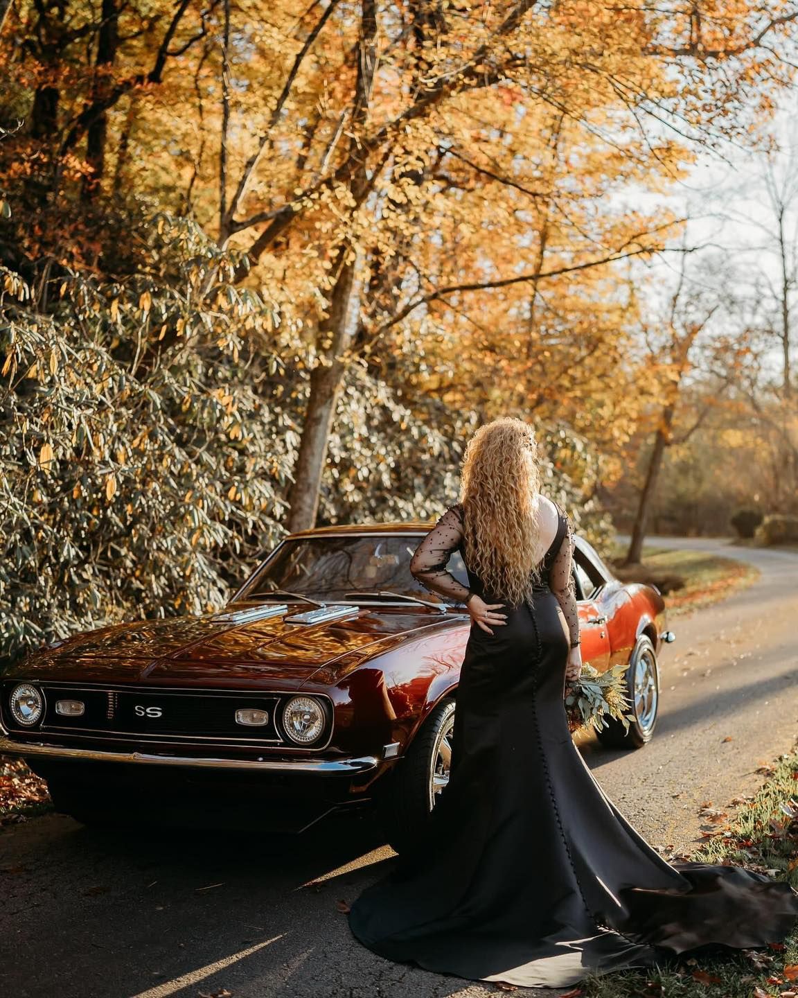 A classic red Camaro parked on an autumn road with golden foliage creating a romantic fall scene.