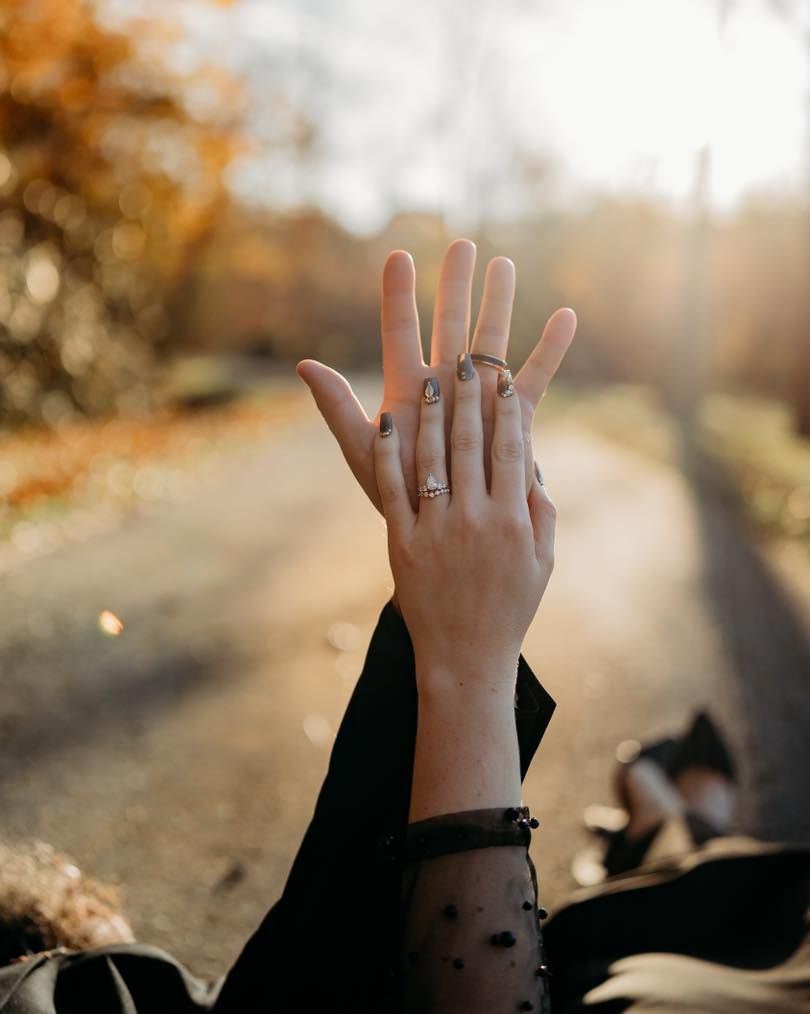 A hand with an engagement ring held up against a sunlit autumn background.
