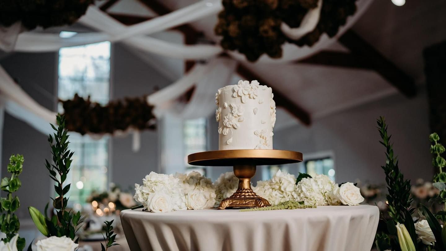 An elegant white wedding cake decorated with flowers sits on a wooden stand at a reception.