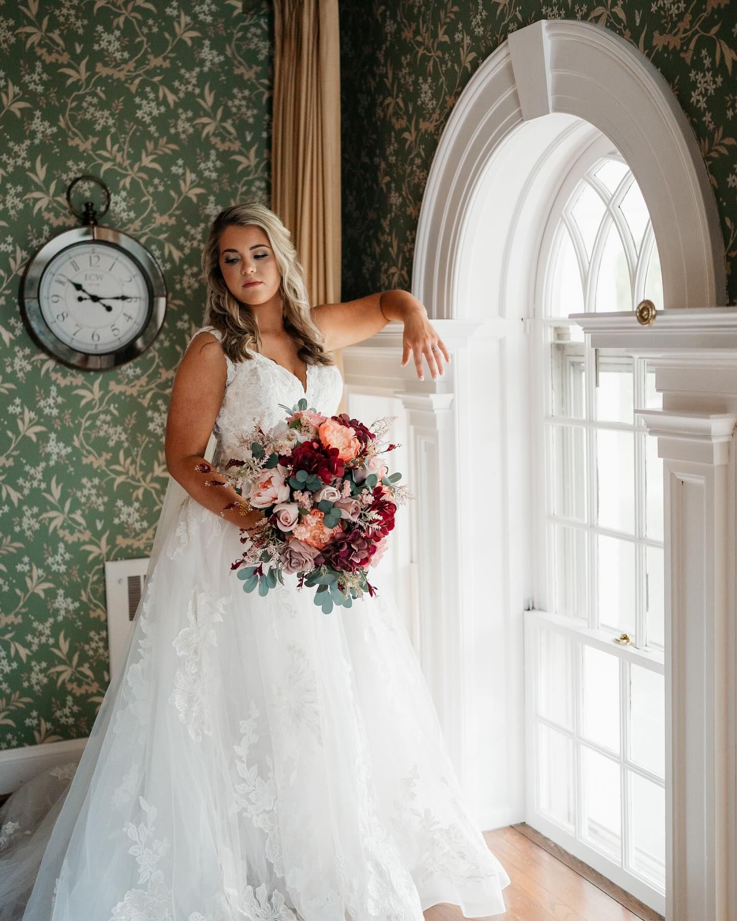 Beautiful bridal portrait with colorful bouquet against green floral wallpaper and arched window.