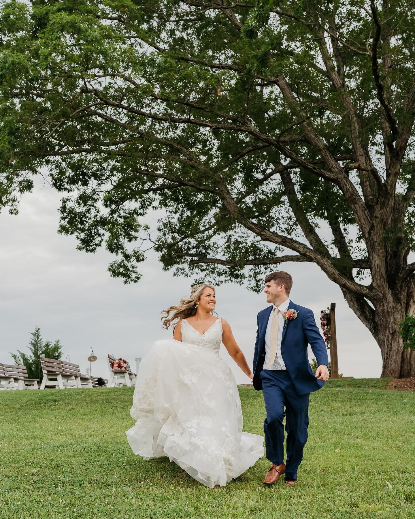 Joyful wedding couple walking on grass beneath large tree with flowing white dress.