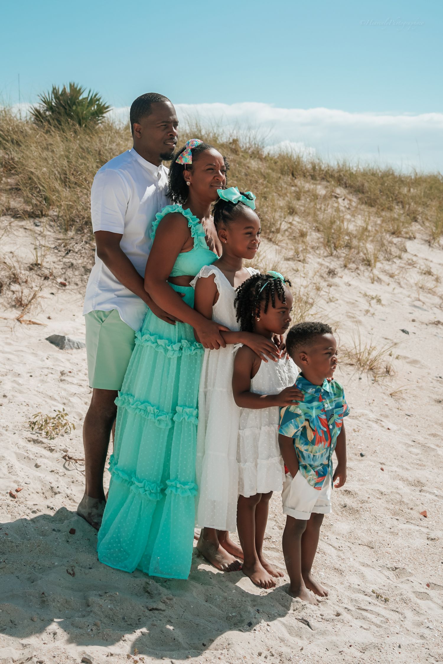 Family members embrace while sitting together on sandy beach dunes under blue skies.