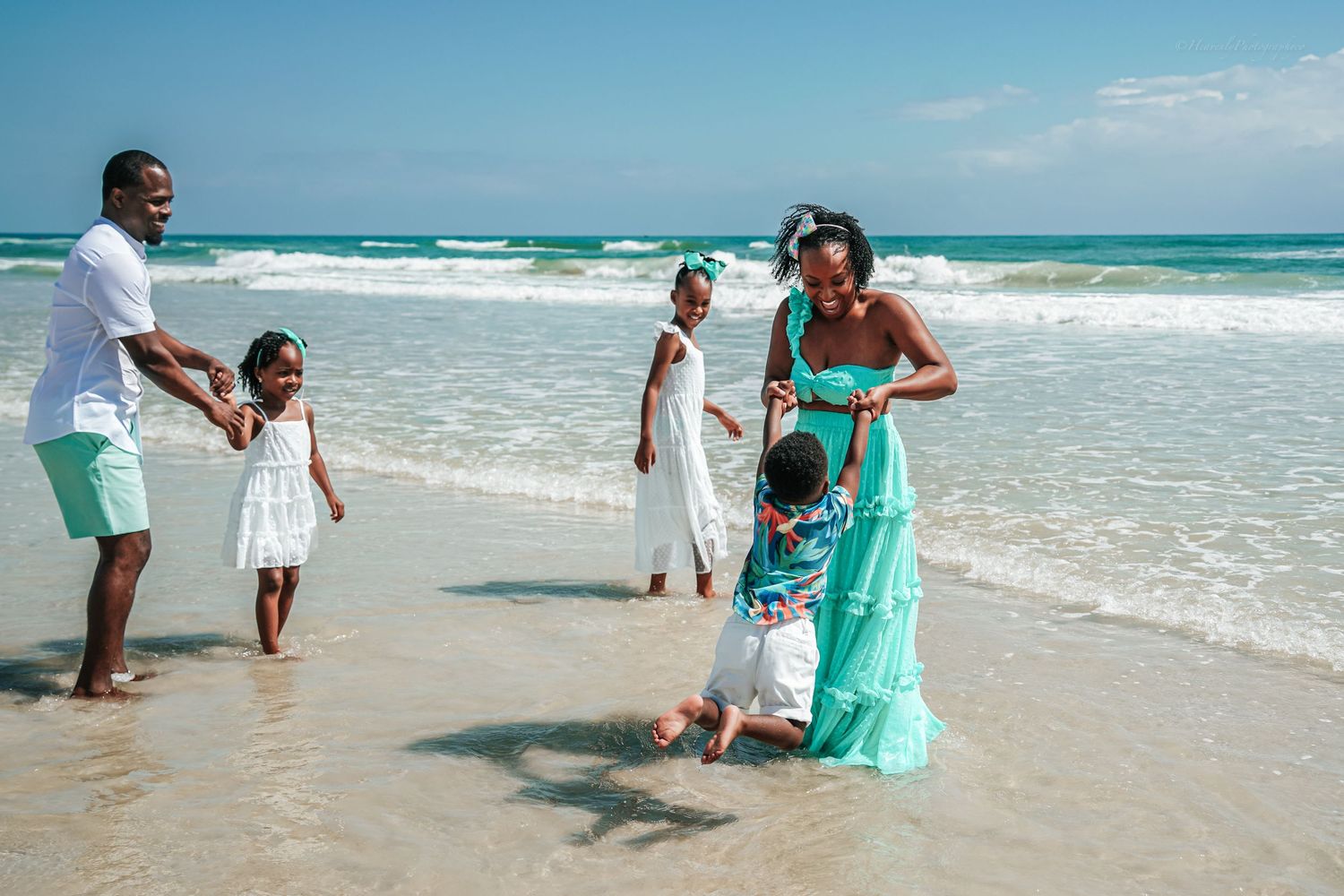 Family members play and splash in shallow ocean waters on sunny beach day.