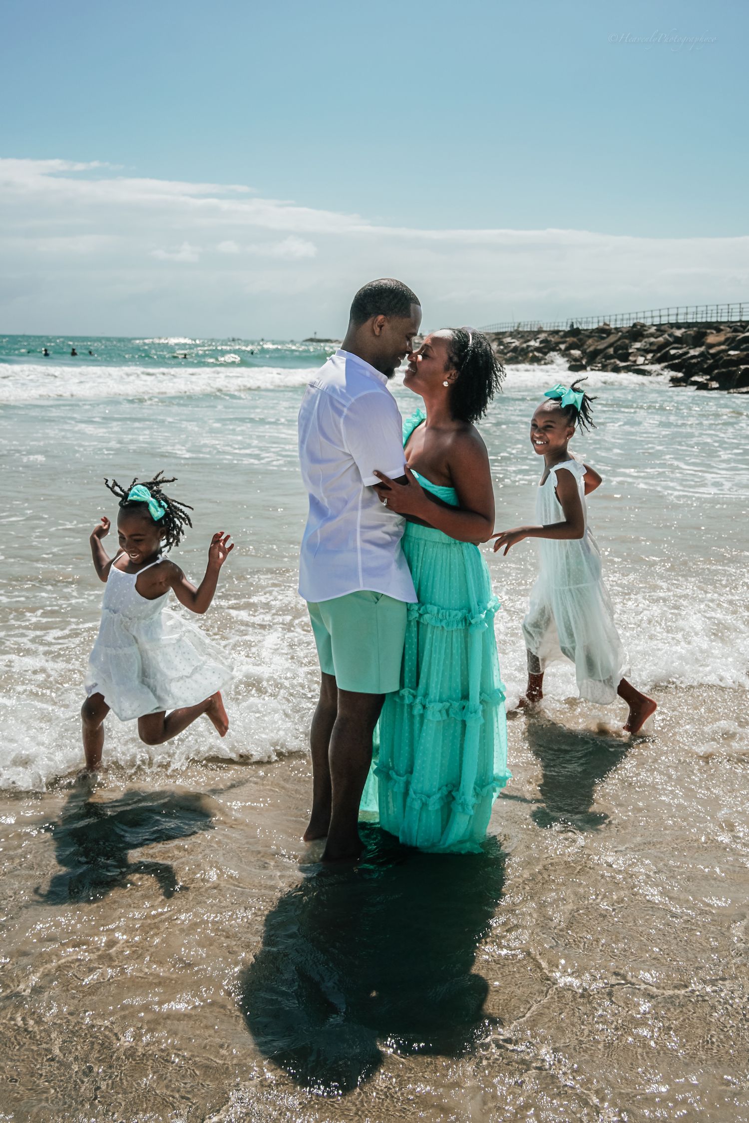 Family wearing white and turquoise outfits playing together in ocean waves on sunny beach day.
