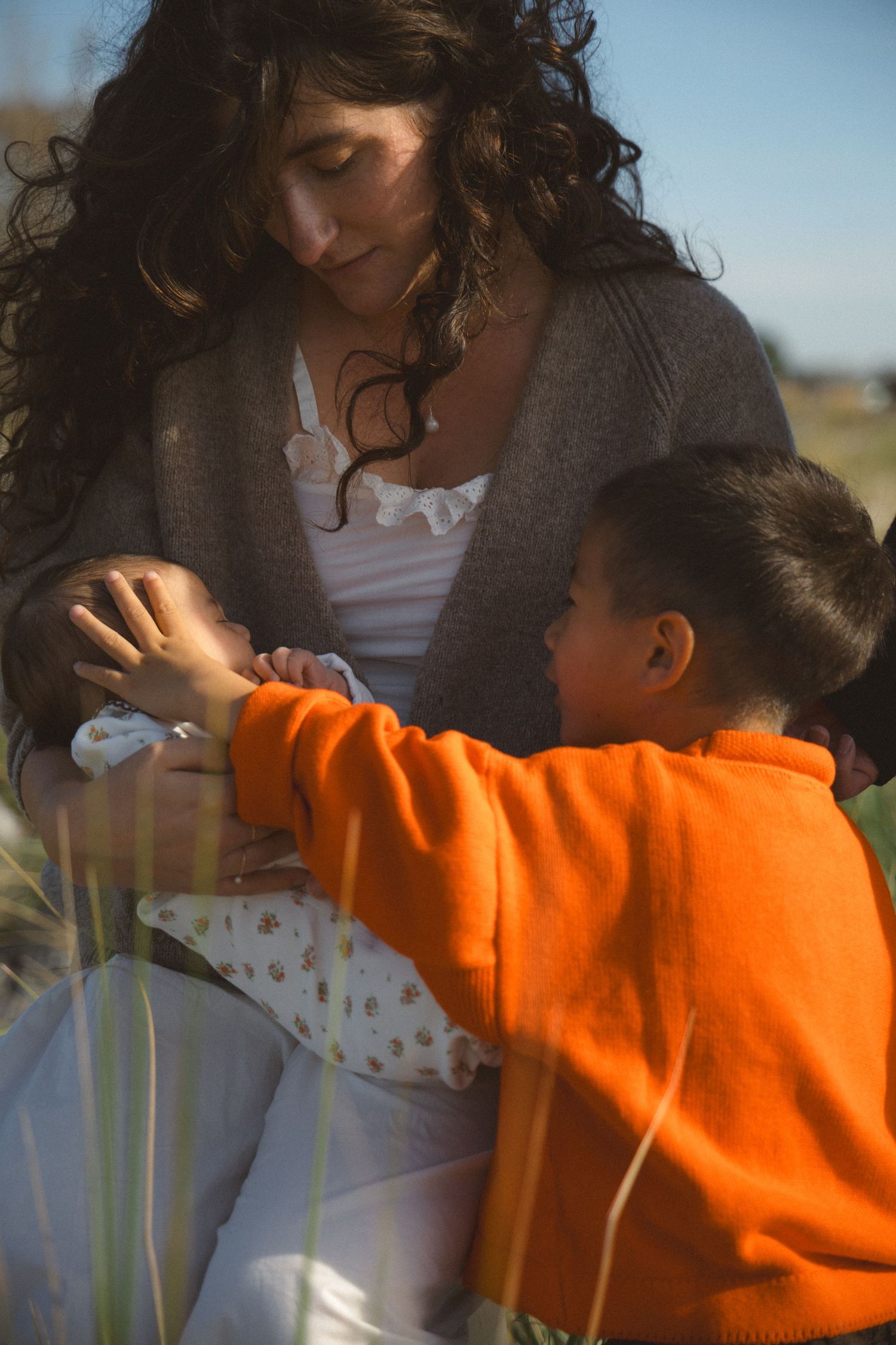 Toddler in orange sweater touching his newborn baby sister gently while mom holds her and looks down at them during a family photoshoot.