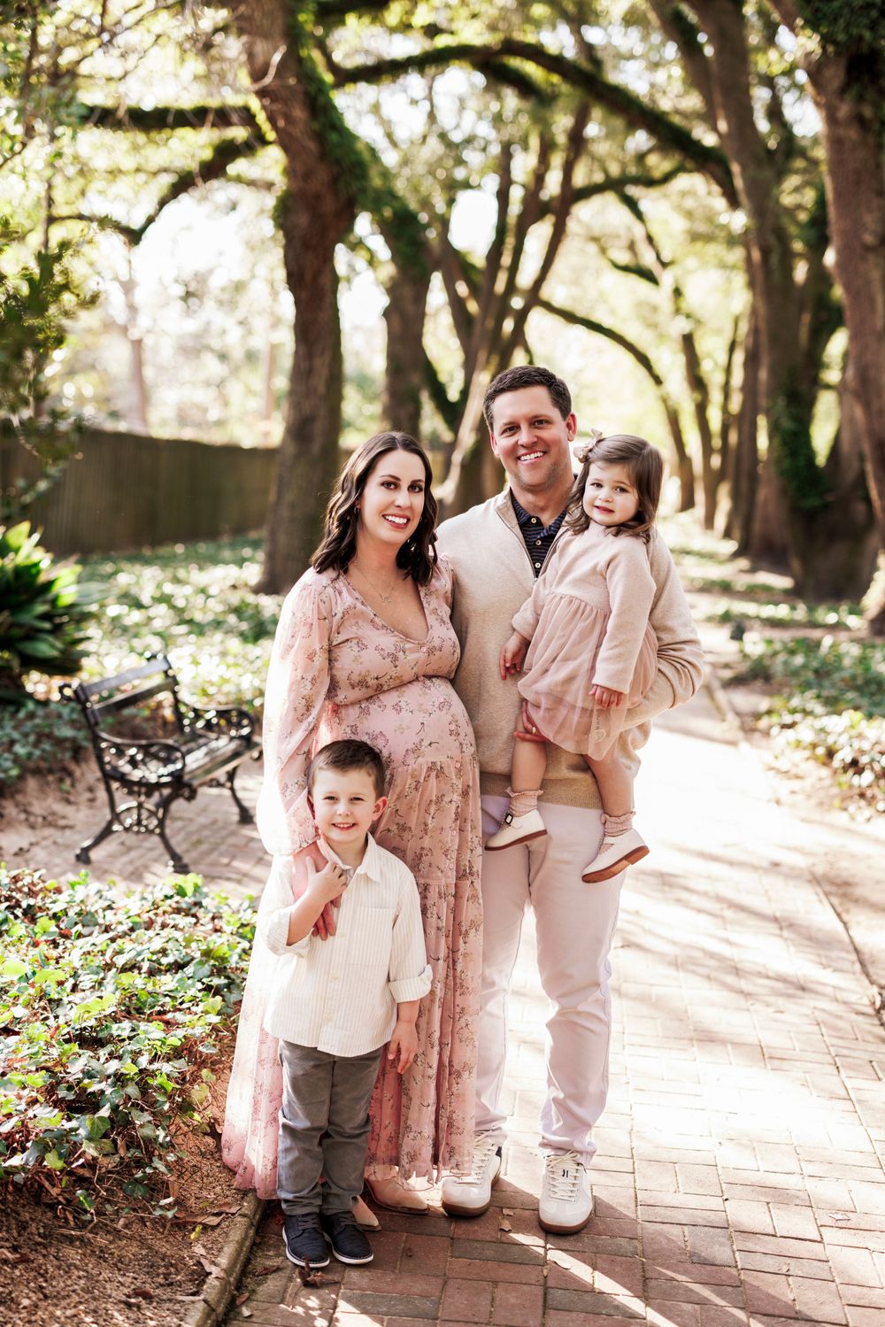Expecting family poses together on brick pathway surrounded by lush trees and greenery in outdoor park setting.