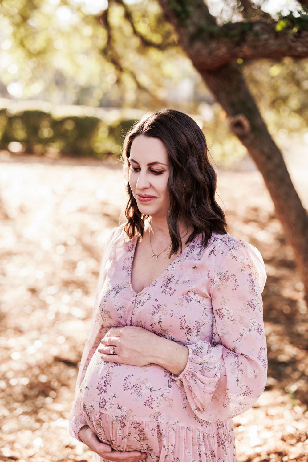 Expectant mother in flowing pink floral dress cradles her baby bump during outdoor maternity photoshoot in golden sunlight.