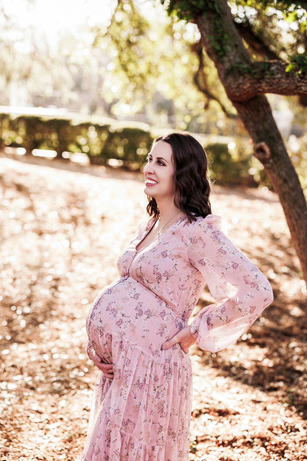 A pregnant woman in a pink floral dress poses outdoors in a sunlit wooded area during an autumn photo session.