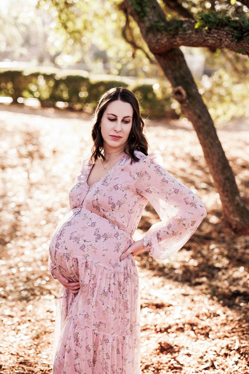 Beautiful maternity photo outdoors showing expecting mother in flowing pink floral dress standing under tree branches.