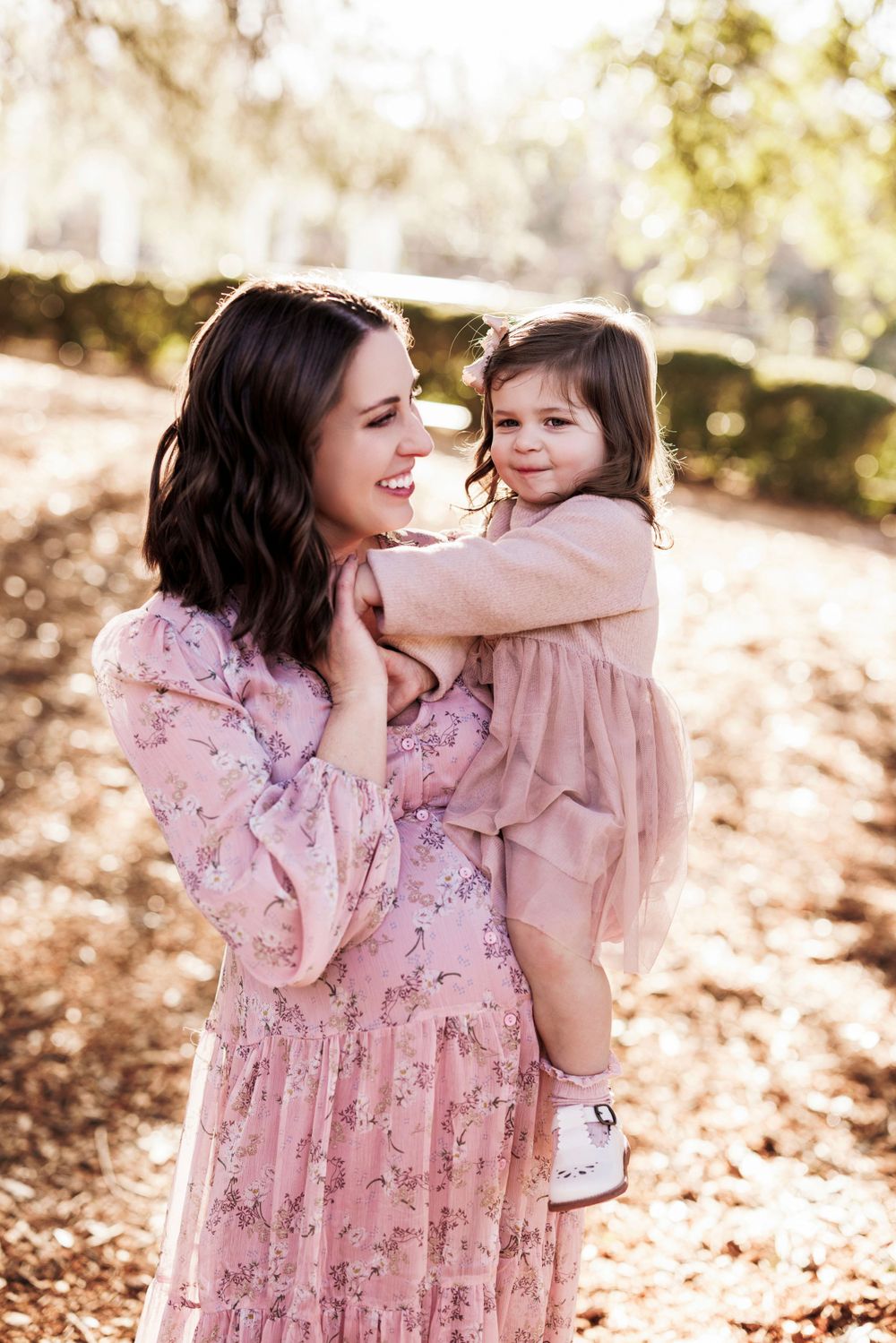 A mother and daughter in matching pink dresses share a tender moment outdoors during an autumn photoshoot.
