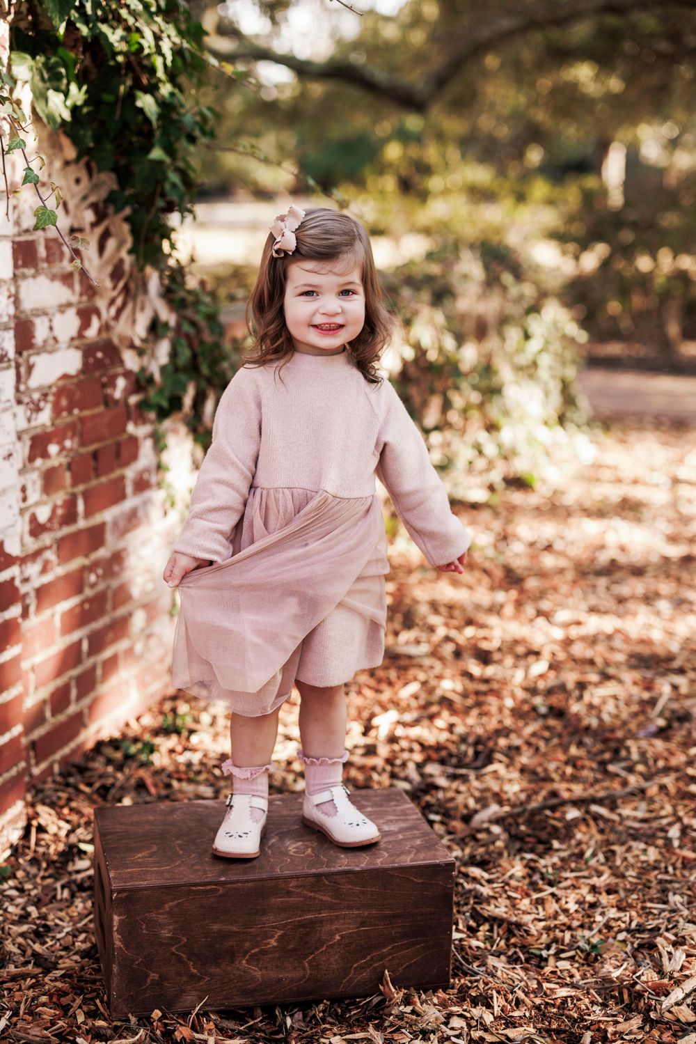 Child in pink dress and white shoes stands on wooden box by brick wall surrounded by autumn leaves.