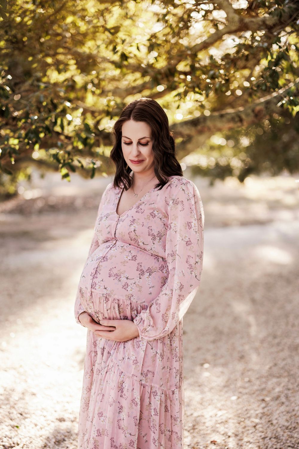 An expectant mother in a flowing pink floral dress cradles her baby bump while standing in dappled sunlight outdoors.