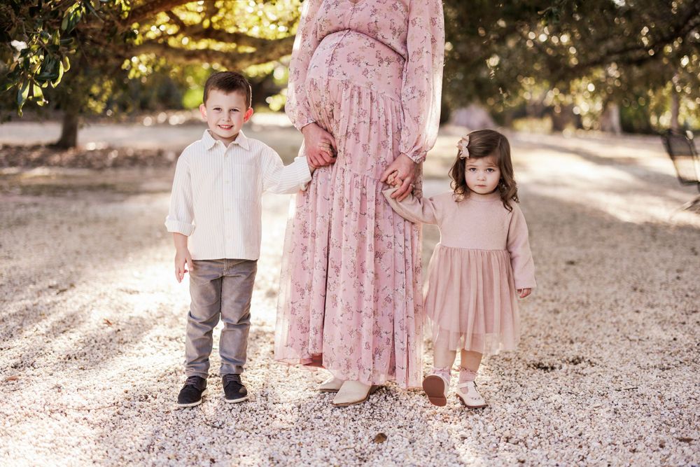 Family members in coordinated outfits stand together on a sunlit path amongst trees wearing pink and neutral tones.