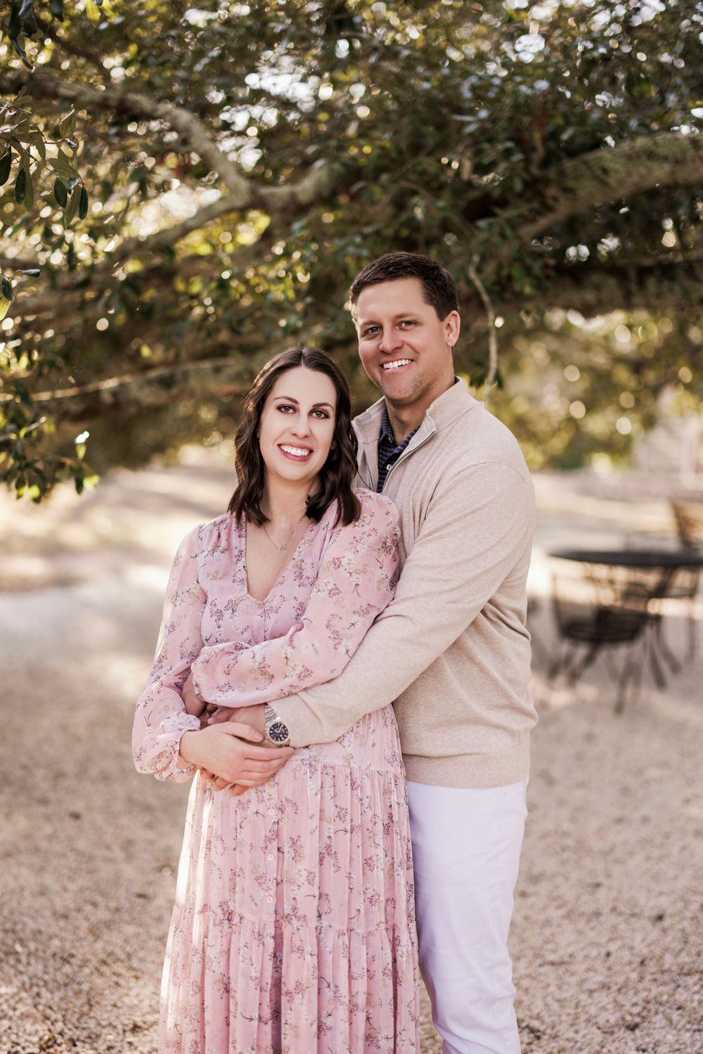 Couple embraces outdoors in romantic springtime setting, wearing a pink floral dress and beige sweater in natural lighting.