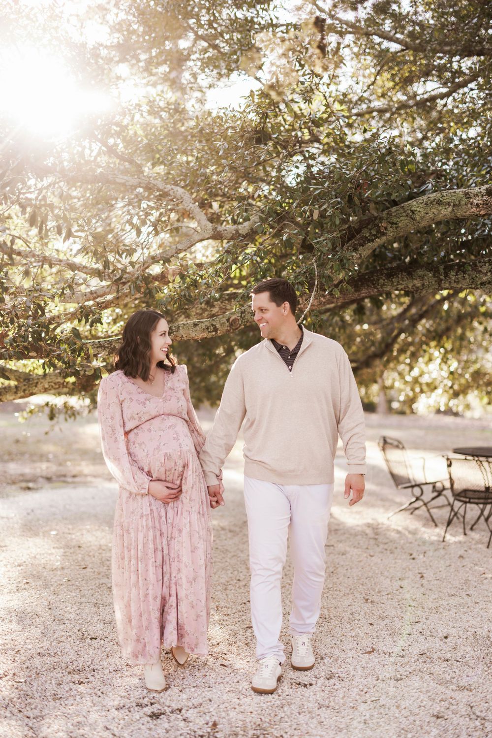 A couple walks hand in hand on a sunlit path under blossoming trees during a romantic maternity photo session.