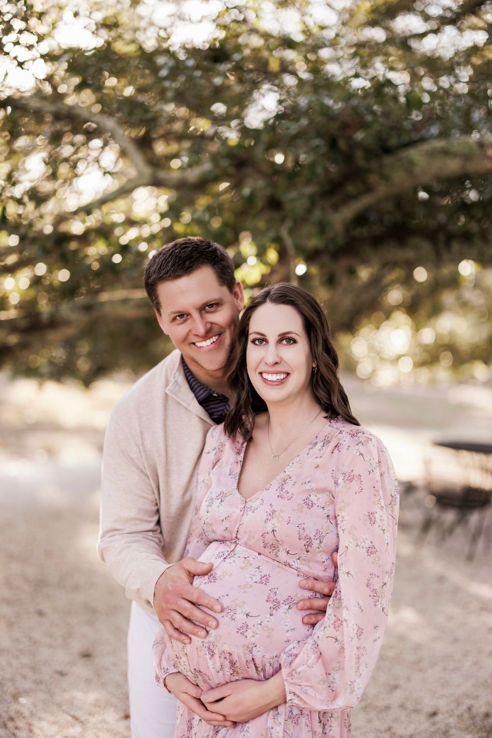 A couple embraces outdoors during maternity photoshoot at sunset with soft bokeh lighting through trees.