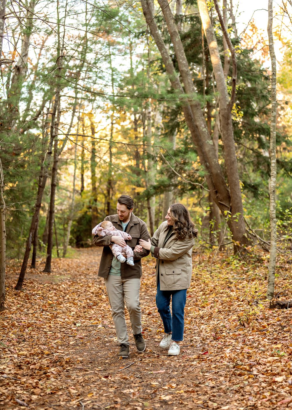 A couple walking with their baby on a leaf-covered trail through an autumn forest during golden hour.