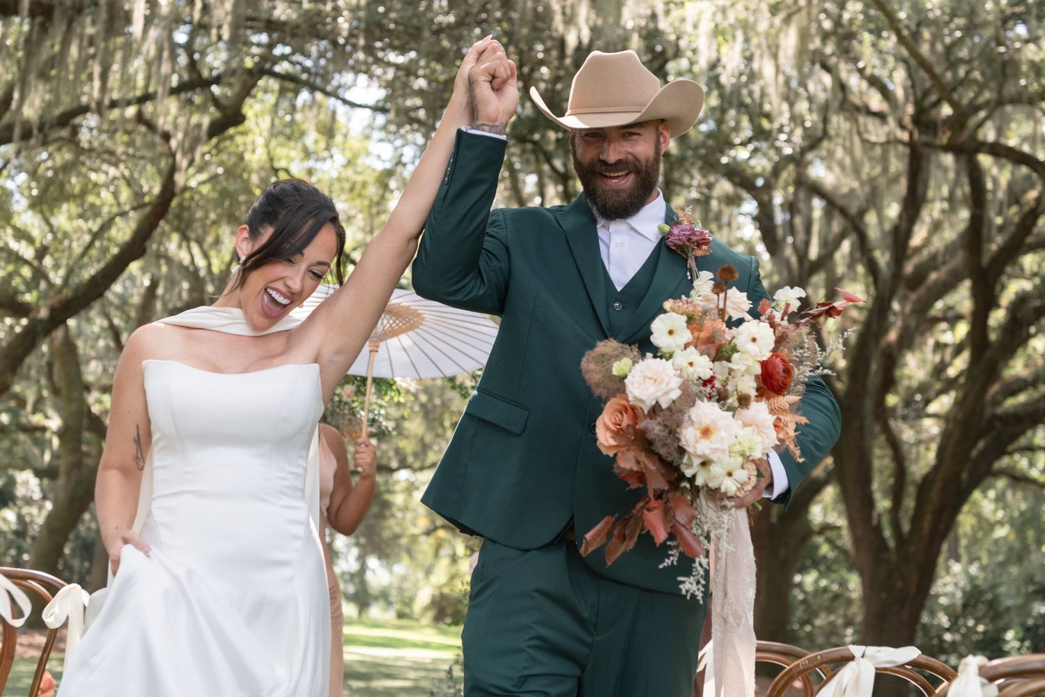 A romantic outdoor wedding celebration with a joyful couple holding hands and a beautiful bouquet in a tree-lined setting.