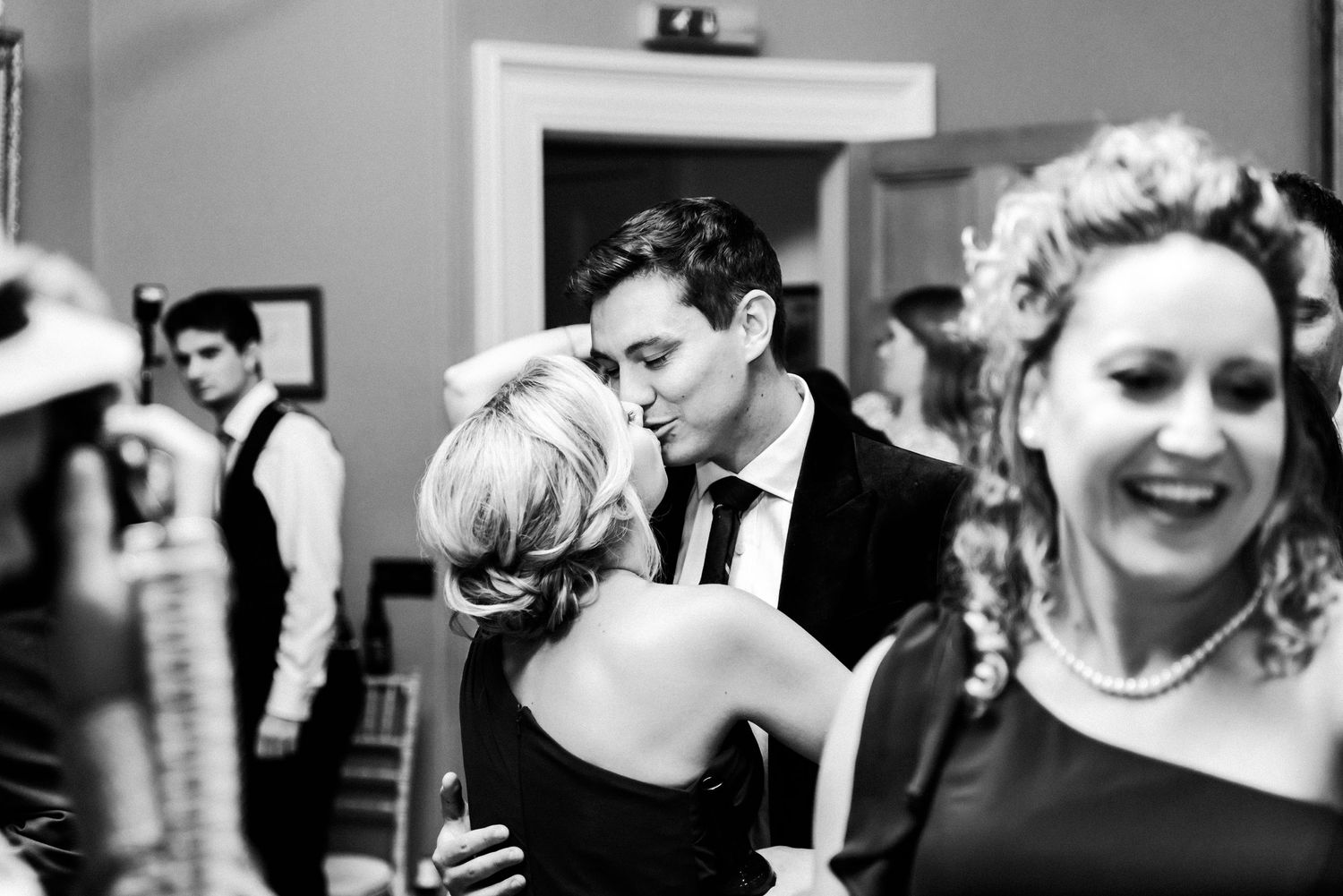 Black and white photo of guests dancing and laughing at a wedding party in an indoor venue.