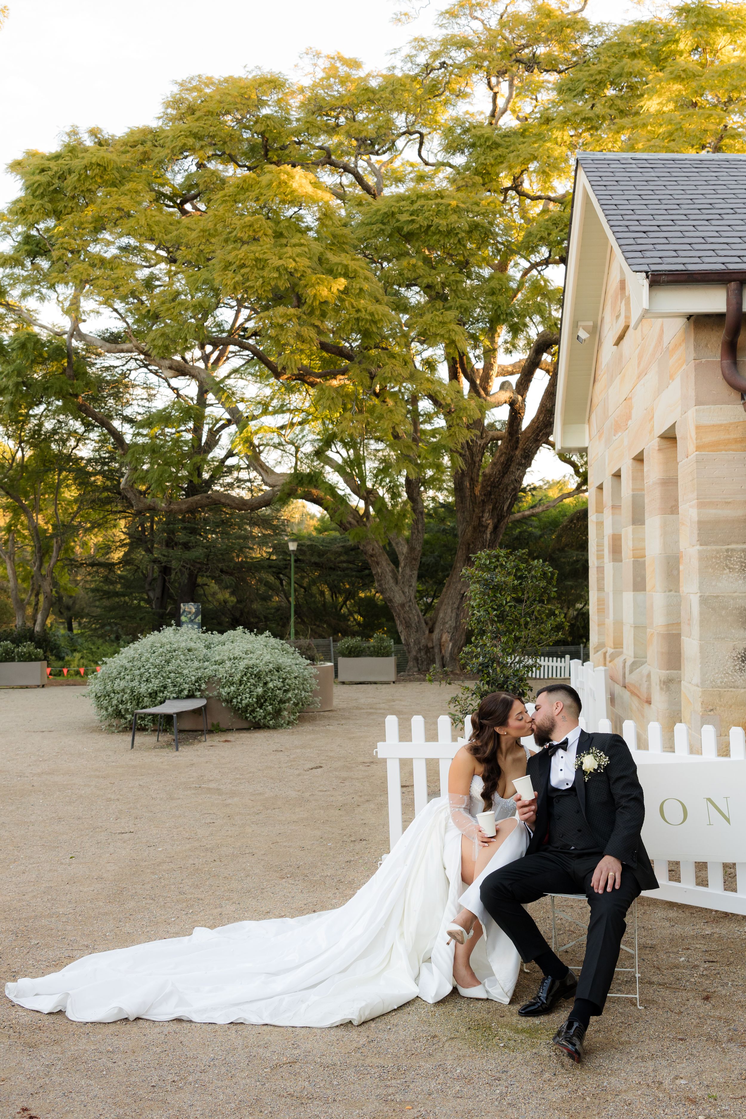 Bride and groom kissing beside white picket fence with large tree in garden setting.
