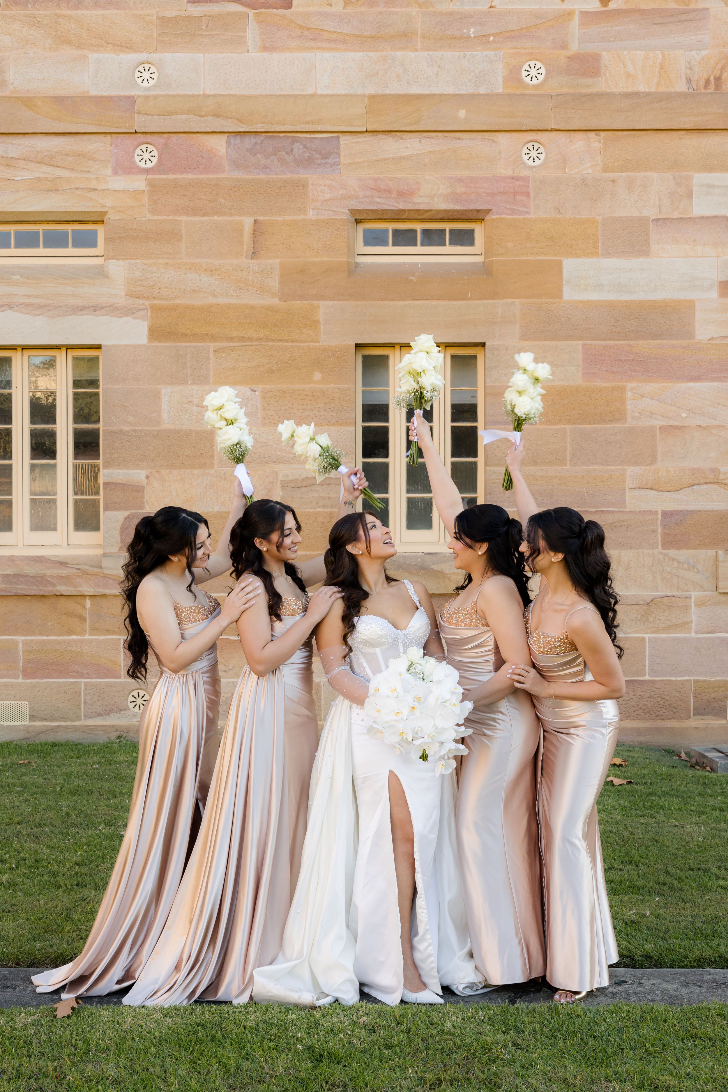 Bride with bridesmaids in champagne satin gowns celebrate joyfully raising bouquets against a sandstone wall.