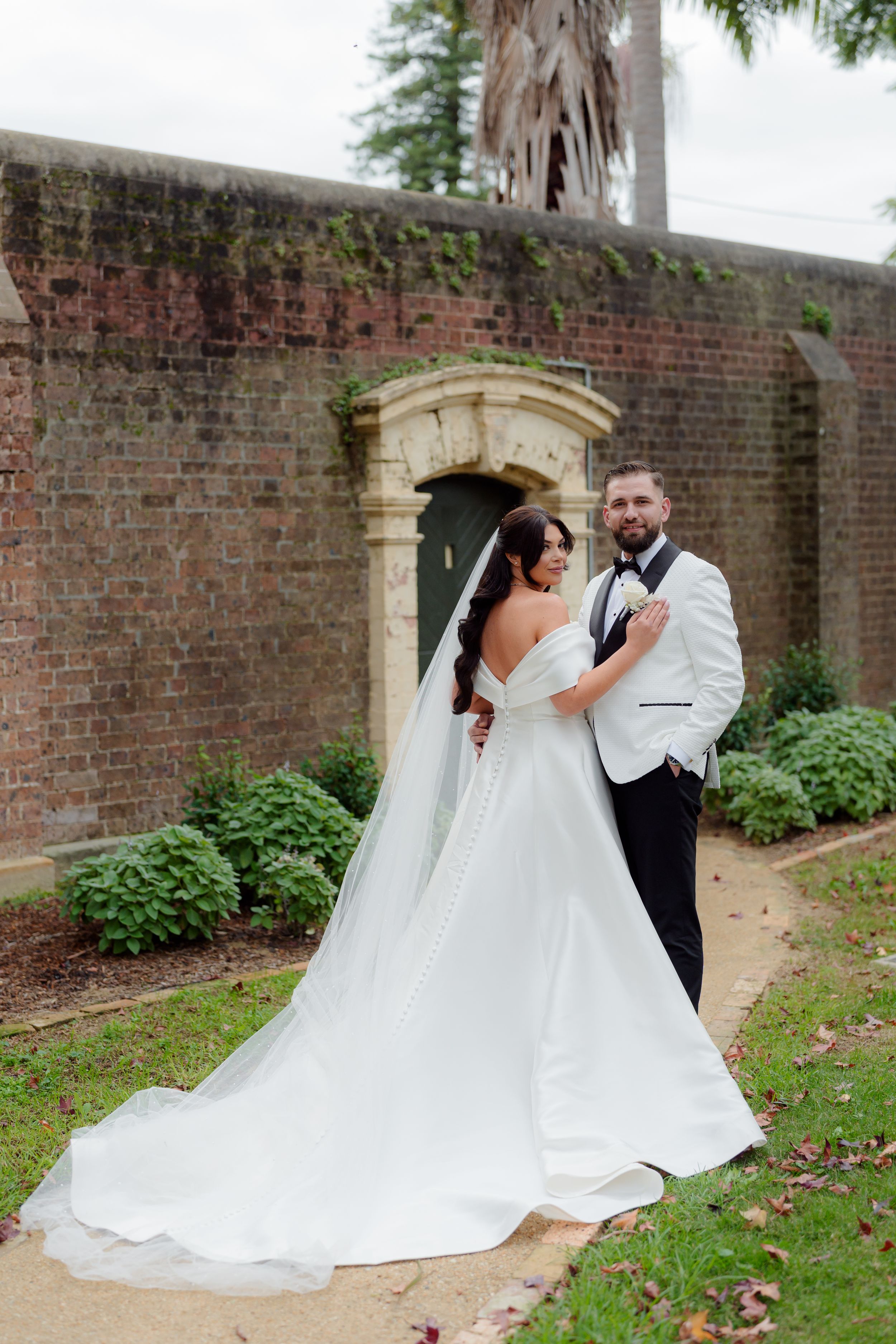 Bride and groom embracing before historic brick wall with stone archway in garden.